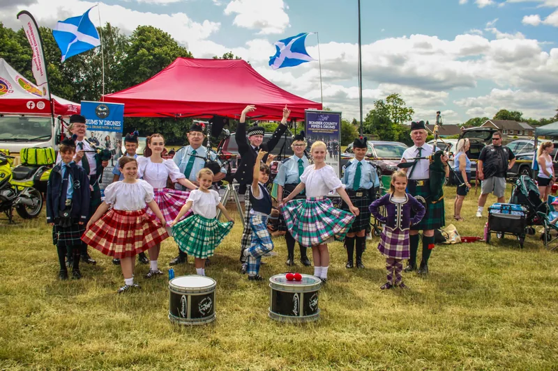 Highland dancers and young band members with drums in front of a red gazebo and Saltire flags