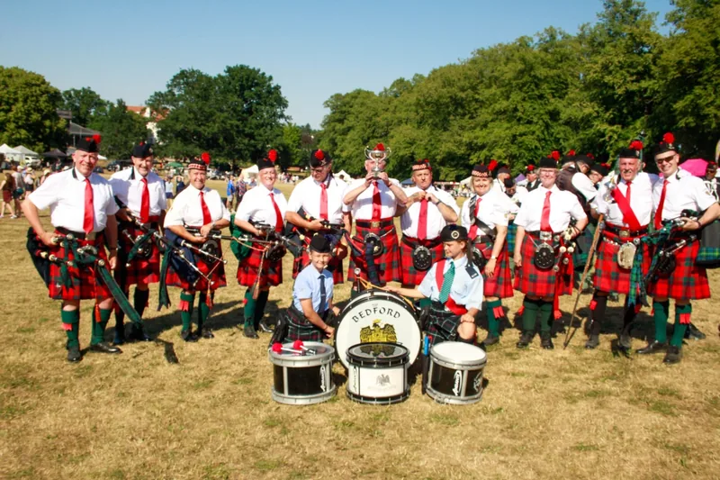 The full band in red tartan lined up on a park field with their drums